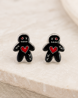 A pair of black voodoo doll stud earrings with red hearts and white stitched details, displayed on a light beige stone surface.