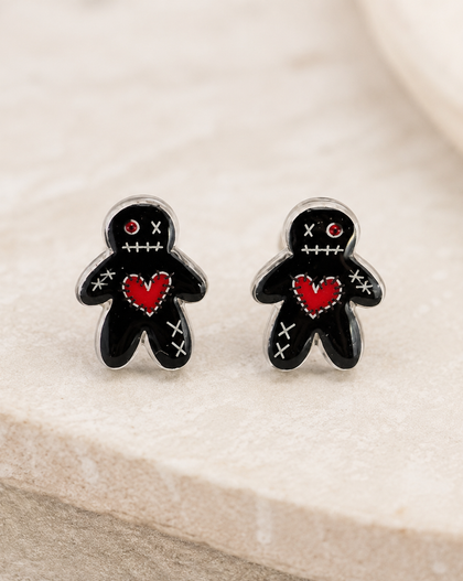 A pair of black voodoo doll stud earrings with red hearts and white stitched details, displayed on a light beige stone surface.
