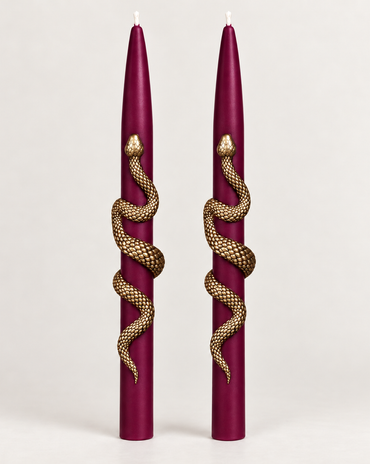 Set of two burgundy taper candles wrapped with gold snake accents on a neutral background.