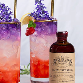 Two cocktails with ice and garnishes, a bottle of El Guapo Cajun Grenadine, and strawberries on a light background.