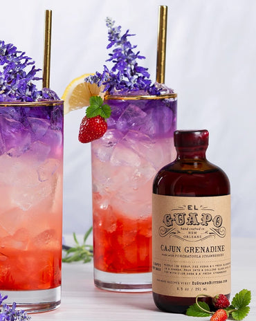 Two cocktails with ice and garnishes, a bottle of El Guapo Cajun Grenadine, and strawberries on a light background.