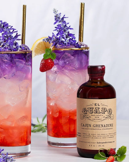 Two cocktails with ice and garnishes, a bottle of El Guapo Cajun Grenadine, and strawberries on a light background.