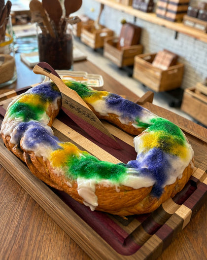 A king cake decorated with white icing and colorful green, purple, and yellow frosting, resting on a wooden cutting board.