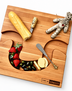NOLA Boards wooden charcuterie board styled with cheese, strawberries, grapes, serving utensils, and a voodoo doll toothpick holder.