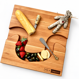 NOLA Boards wooden charcuterie board styled with cheese, strawberries, grapes, serving utensils, and a voodoo doll toothpick holder.