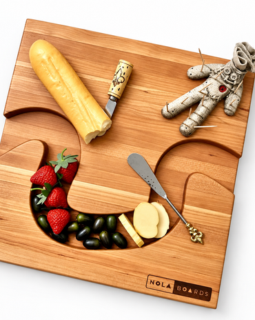 NOLA Boards wooden charcuterie board styled with cheese, strawberries, grapes, serving utensils, and a voodoo doll toothpick holder.