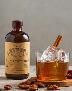 Bottle of El Guapo Sweet Potato Syrup with a glass of iced drink and pecans on a wooden surface.