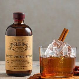Bottle of El Guapo Sweet Potato Syrup with a glass of iced drink and pecans on a wooden surface.