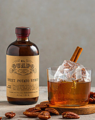 Bottle of El Guapo Sweet Potato Syrup with a glass of iced drink and pecans on a wooden surface.