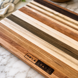 Wooden cutting board with striped pattern on a marble surface, featuring 'Nola Boards' branding.