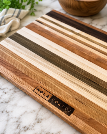 Wooden cutting board with striped pattern on a marble surface, featuring 'Nola Boards' branding.
