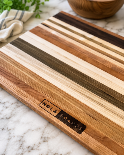 Wooden cutting board with striped pattern on a marble surface, featuring 'Nola Boards' branding.